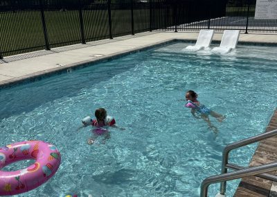 "Two young girls swimming in an outdoor pool at Homestead RV Community, with a unicorn and cupcake-print pool float nearby and a shaded lawn area behind the pool fence.
