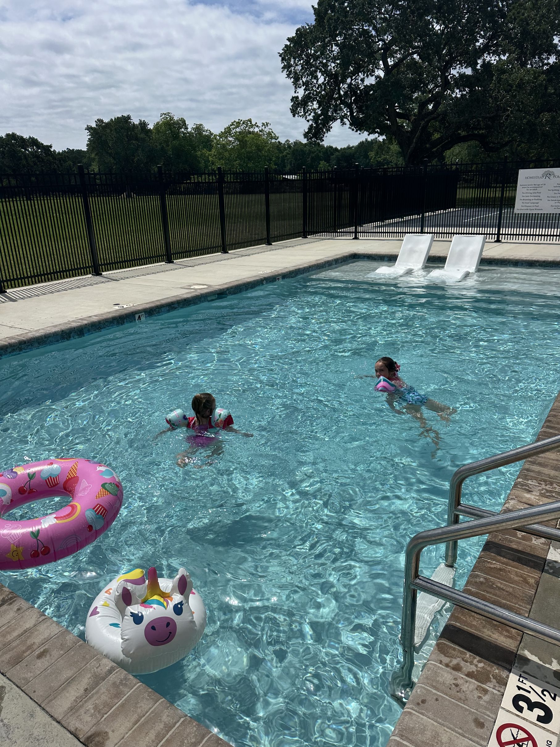 "Two young girls swimming in an outdoor pool at Homestead RV Community, with a unicorn and cupcake-print pool float nearby and a shaded lawn area behind the pool fence.
