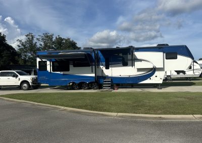 A blue and white Luxe fifth-wheel RV with awning extended, parked beside a white dually pickup truck on a paved site at Homestead RV Community, under a partly cloudy sky.
